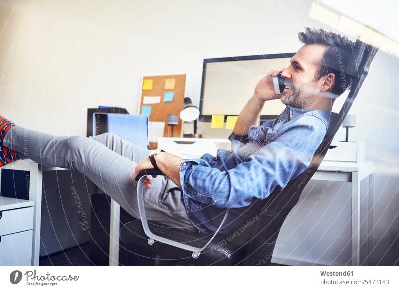 Entspannter Mann sitzt am Schreibtisch im Büro und telefoniert mit dem Handy telefonieren anrufen Anruf telephonieren sitzen sitzend Mobiltelefon Handies Handys