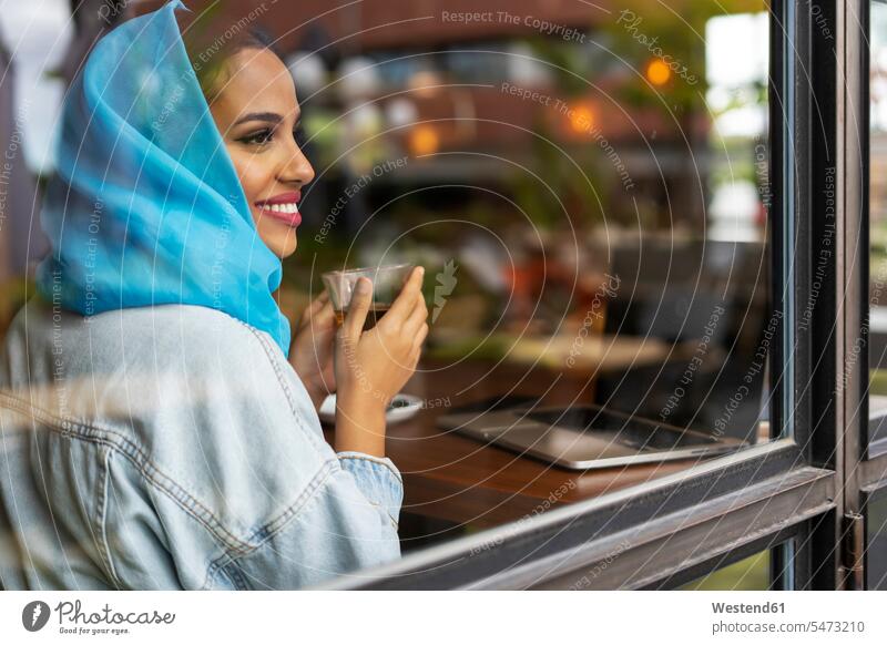 Junge Frau mit türkisfarbenem Hidschab, die in einem Café Tee trinkt und durch ein Fenster schaut Kopftücher Rechner Laptops Notebook Notebooks sitzend sitzt