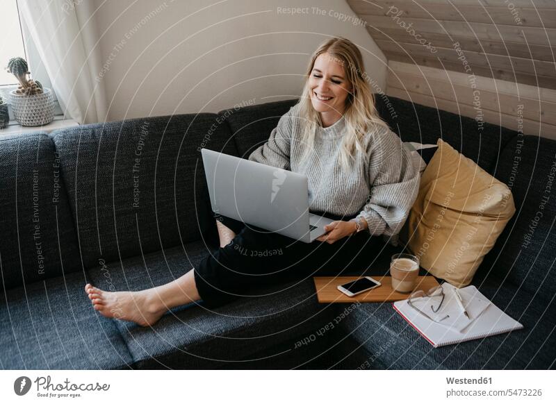Lächelnder Freiberufler mit Laptop, während er zu Hause mit Getränk und Dokument auf dem Sofa sitzt Farbaufnahme Farbe Farbfoto Farbphoto Innenaufnahme
