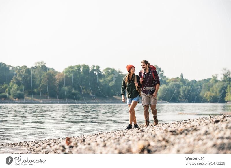 Lächelndes junges Paar mit Rucksäcken beim Spaziergang am Flussufer Rucksack Fluesse Fluß Flüsse Pärchen Paare Partnerschaft lächeln gehen gehend geht Gewässer