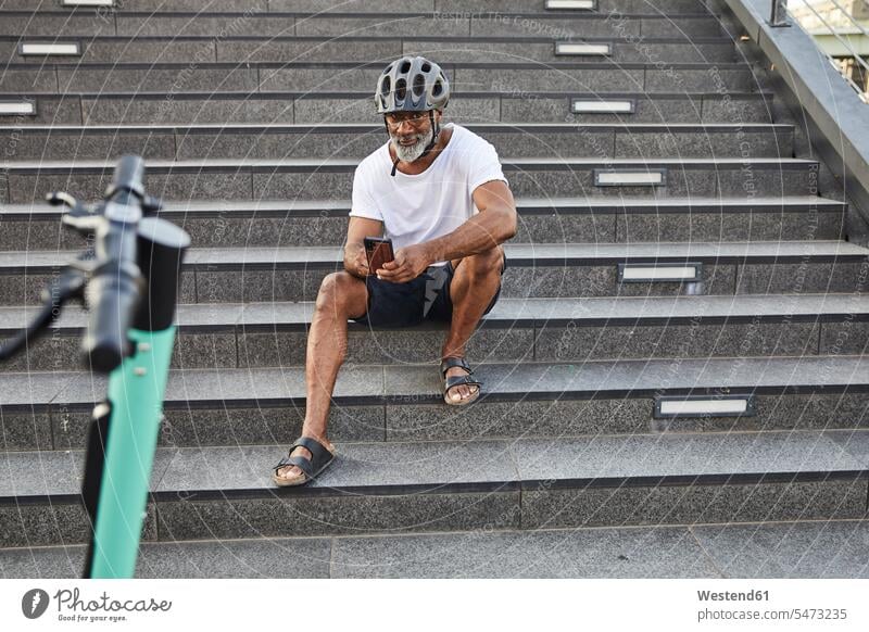 Lächelnder reifer Mann mit E-Scooter, der auf einer Treppe sitzt und ein Mobiltelefon benutzt Leute Menschen People Person Personen gemischtrassig 1 Ein