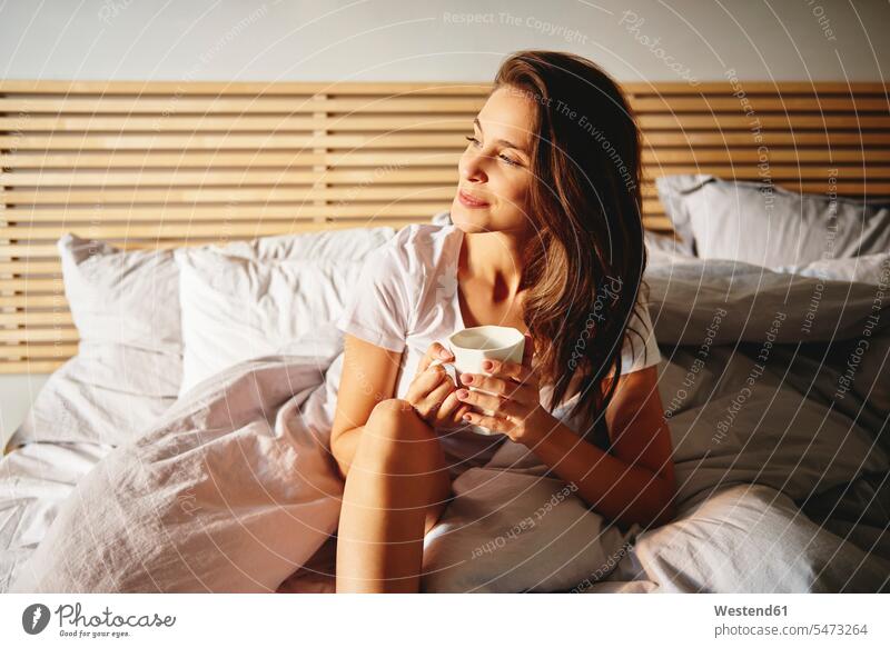 Porträt einer lächelnden jungen Frau, die mit einer Tasse Kaffee auf dem Bett sitzt und in die Ferne schaut Betten schauen sehend sitzen sitzend weiblich Frauen