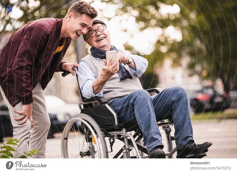 Lachender älterer Mann im Rollstuhl und sein erwachsener Enkel schauen gemeinsam auf Smartphone und haben Spaß Generation Handies Handys Mobiltelefon
