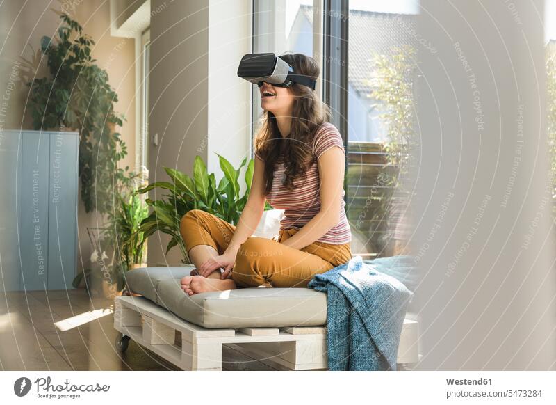 Glückliche junge Frau sitzt am Fenster zu Hause mit VR-Brille virtuell Virtualität weiblich Frauen Zuhause daheim glücklich glücklich sein glücklichsein Brillen