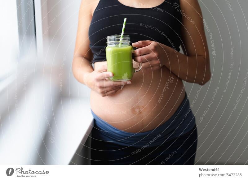 Junge schwangere Frau trinkt frischen und gesunden grünen Smoothie Gläser Trinkglas Trinkgläser Strohhalme Trinkhalm Trinkhalme stehend steht daheim zu Hause