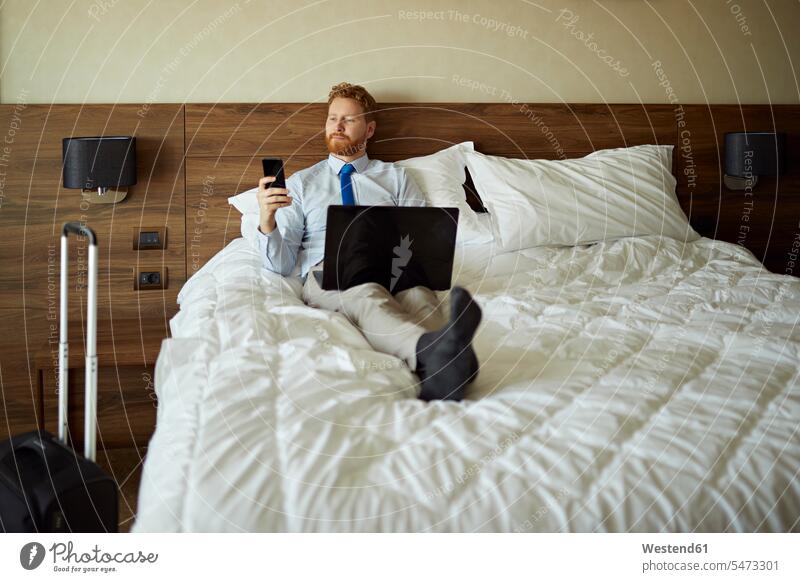 Geschäftsmann liegt auf dem Bett im Hotelzimmer und benutzt Laptop und Mobiltelefon Job Berufe Berufstätigkeit Beschäftigung Jobs geschäftlich Geschäftsleben