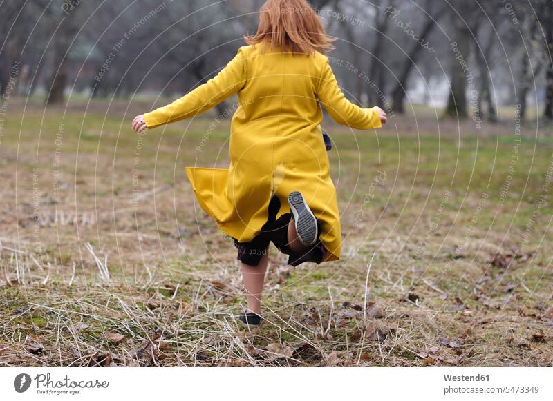 Rückansicht einer Frau in gelbem Mantel, die im Park auf Gras läuft Farbaufnahme Farbe Farbfoto Farbphoto Außenaufnahme außen draußen im Freien Tag