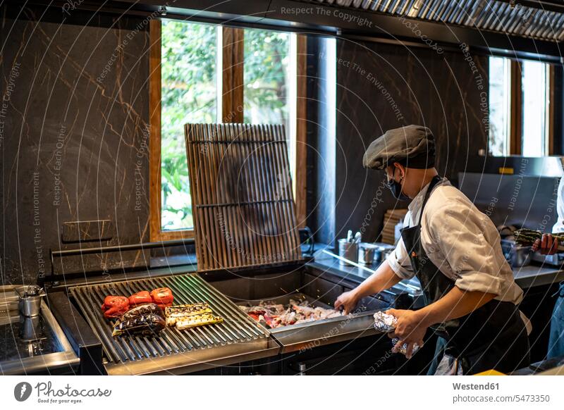 Chefkoch mit Gesichtsschutzmaske bei der Zubereitung von gegrilltem Gemüse in der Restaurantküche Arbeitsbekleidung drinnen Innenaufnahmen Gastronomie Koeche