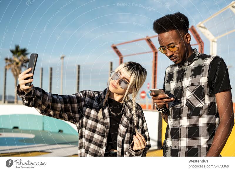 Junge Frau macht Selfie mit ihrem Freund und schaut auf sein Smartphone Leute Menschen People Person Personen Europäisch Kaukasier kaukasisch Afrikanisch