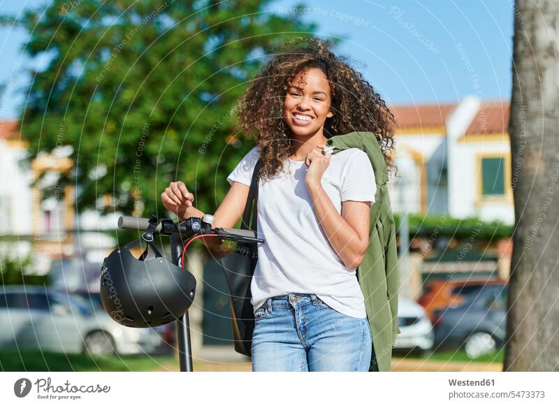 Glückliche junge Frau trägt Jacke, während sie an einem sonnigen Tag mit einem elektrischen Roller steht Farbaufnahme Farbe Farbfoto Farbphoto Außenaufnahme