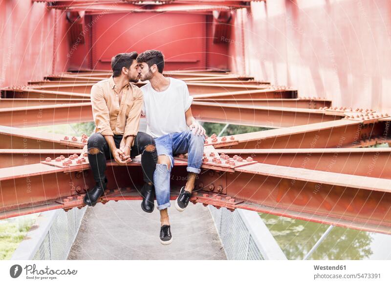 Küssendes junges schwules Paar, das auf dem Stahlträger einer Fußgängerbrücke sitzt sitzen sitzend küssen Kuss Pärchen Paare Partnerschaft Homosexueller Mann