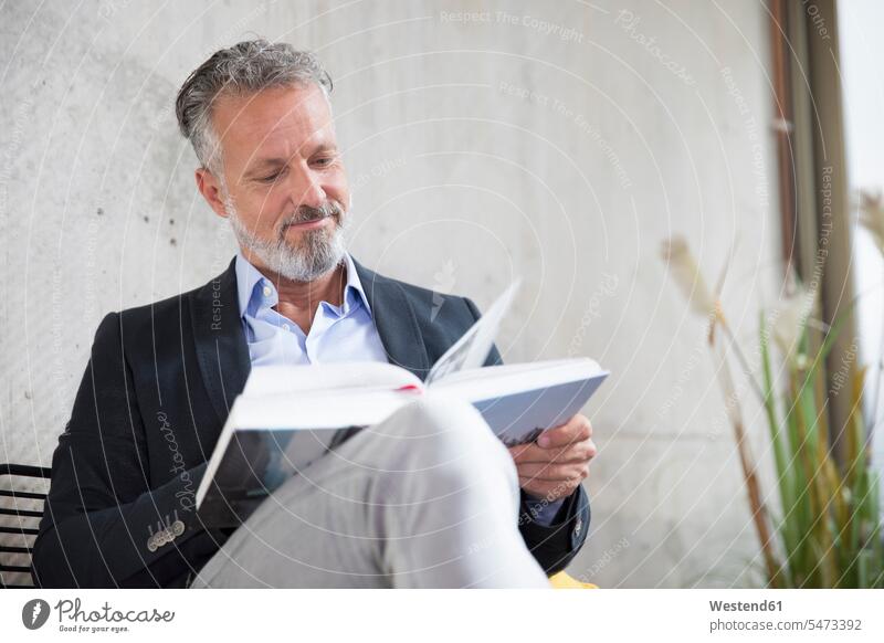 Lächelnder Geschäftsmann sitzt an einer Betonwand und liest ein Buch lesen Lektüre sitzen sitzend Bücher Betonwände Betonwaende Businessmann Businessmänner