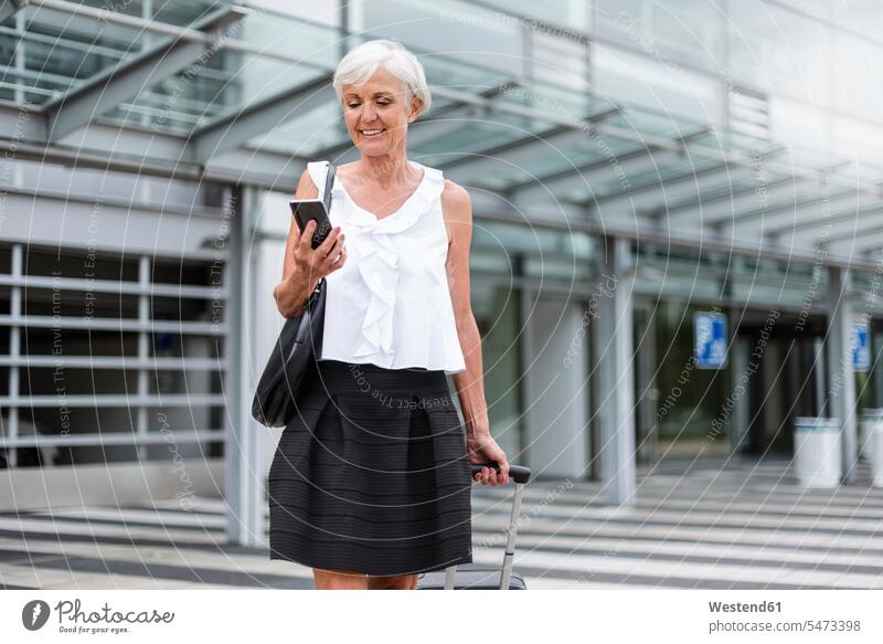 Lächelnde ältere Frau mit Gepäck schaut auf Handy lächeln Seniorin Seniorinnen alt Mobiltelefon Handies Handys Mobiltelefone ansehen weiblich Frauen Reisegepäck
