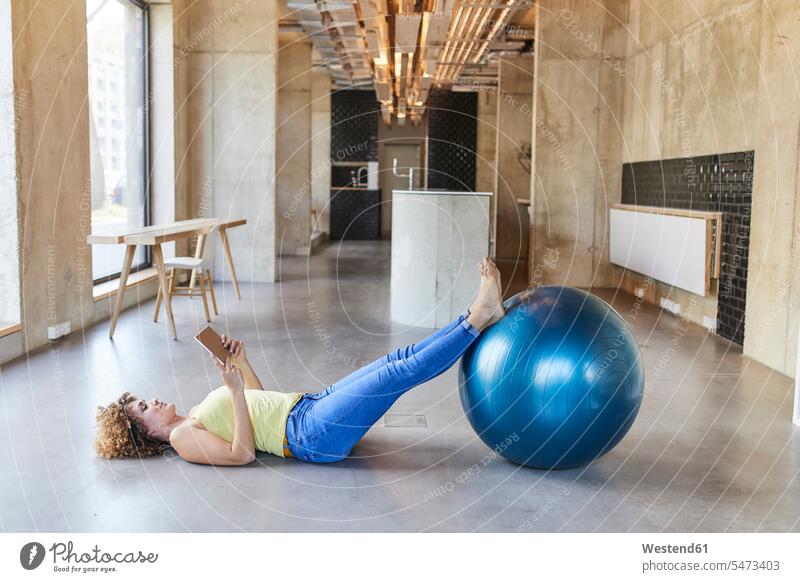 Frau mit Fitness-Ball auf dem Boden liegend mit Tablet in modernen Büro Deutschland Fitnessball Gymnastikball Sitzball Tablet Computer Tablet-PC Tablet PC iPad