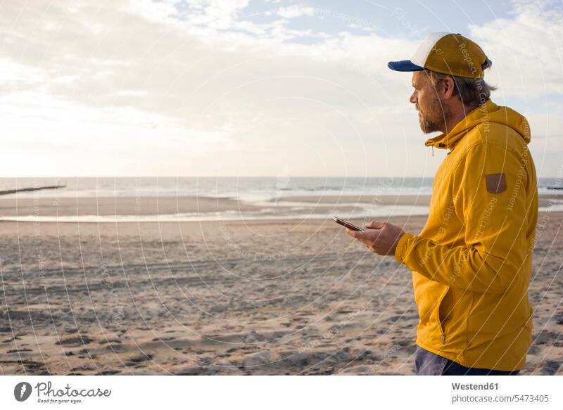 Mann in gelber Jacke, mit Smartphone am Strand benutzen benützen gelbes Jacken iPhone Smartphones Beach Straende Strände Beaches Männer männlich Handy