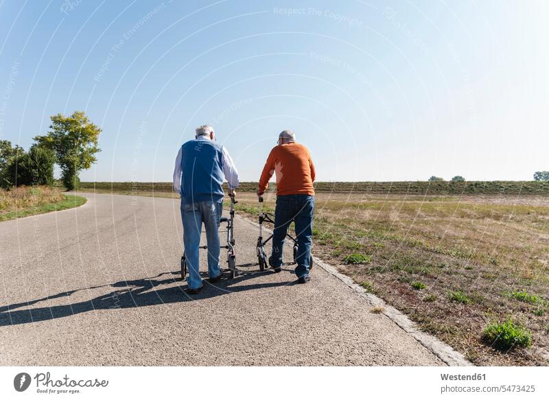 Zwei alte Freunde, die auf einer Landstraße spazieren gehen, mit Rollatoren Europäer Kaukasier Europäisch kaukasisch Beste Freunde Bester Freund zwei Personen 2