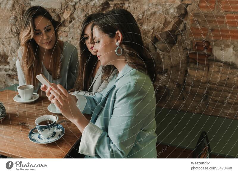 Drei junge Frauen mit einem Mobiltelefon in einem Cafe Spanien Ziegelmauer Ziegelwand Backsteinmauer SMS Textnachricht Backsteinwand Backsteinmauern modisch