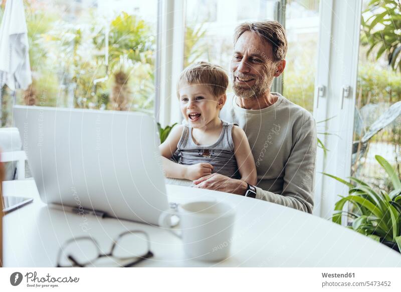 Kleiner Junge sitzt auf dem Schoß seines Vaters und geht von zu Hause zur Arbeit Job Berufe Berufstätigkeit Beschäftigung Jobs Tische Rechner Laptops Notebook