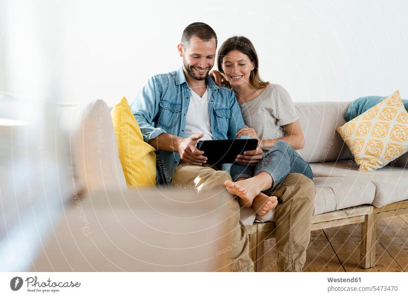 Glückliches Paar zu Hause im modernen Wohnzimmer auf der Couch sitzend, während sie gemeinsam auf ein Tablet schauen Polster Couches Liege Sofas Medium sitzt