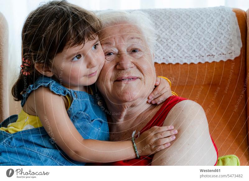 Lächelnde Großmutter umarmt zu Hause ihre Enkelin Stuehle Stühle anfassen Berührung entspannen relaxen knuddeln schmusen sitzend sitzt Arm umlegen Umarmung