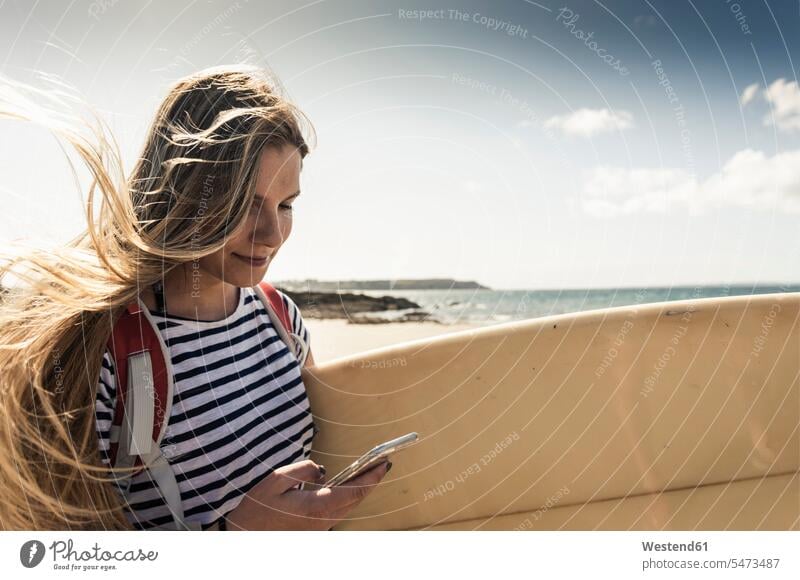 Junge Frau am Strand, trägt Surfbrett, mit Smartphone junge Frau junge Frauen Beach Straende Strände Beaches Surferin Wellenreiterinnen Surferinnen benutzen