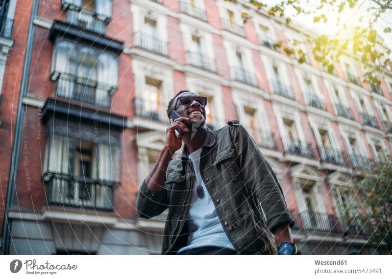 Lächelnder Mann mit Sonnenbrille spricht über Smartphone, während er in der Stadt vor einem Gebäude sitzt Farbaufnahme Farbe Farbfoto Farbphoto Spanien