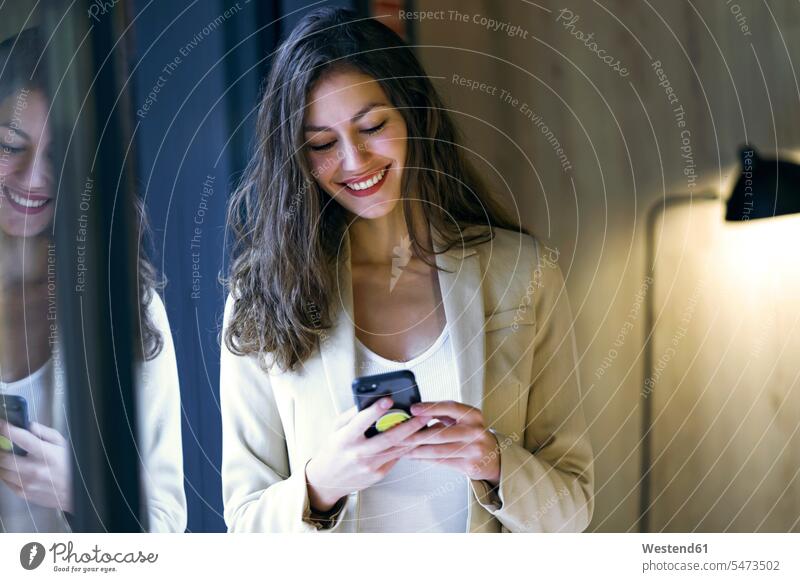 Lächelnde junge Geschäftsfrau überprüft Handy am Fenster Leute Menschen People Person Personen Europäisch Kaukasier kaukasisch 1 Ein ein Mensch eine