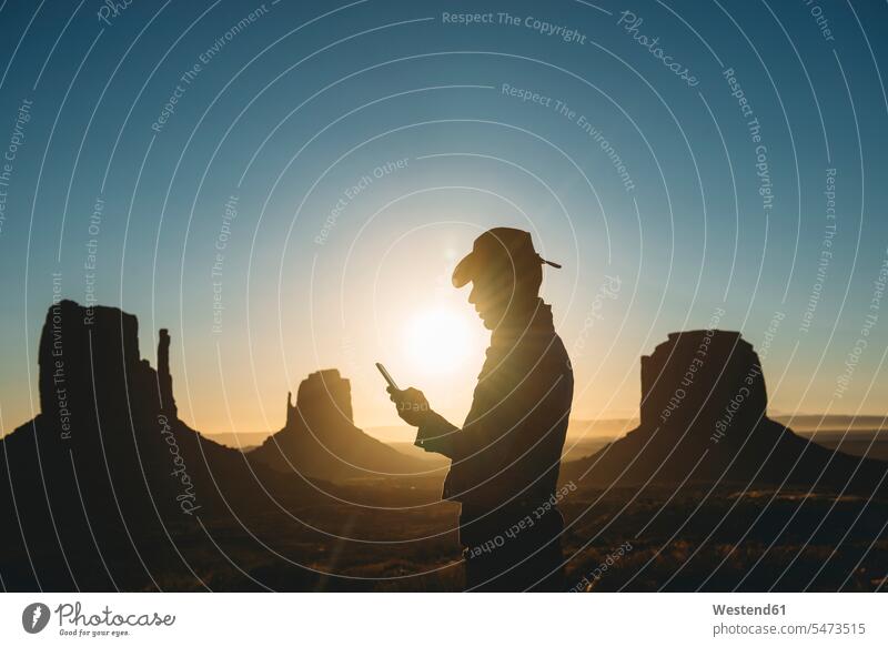 USA, Utah, Monument Valley, Silhouette eines Mannes mit Cowboyhut, der bei Sonnenaufgang auf sein Mobiltelefon schaut Männer männlich Cowboyhuete Cowboyhüte
