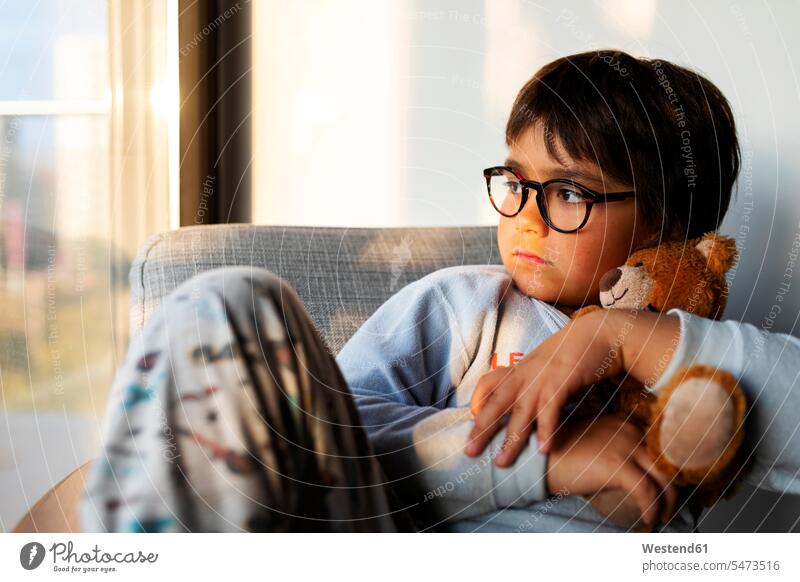 Porträt eines ernsthaften Jungen mit Teddybär, der zu Hause auf einem Sessel sitzt und aus dem Fenster schaut Fensterscheiben Stuehle Stühle Teddies Teddybaer