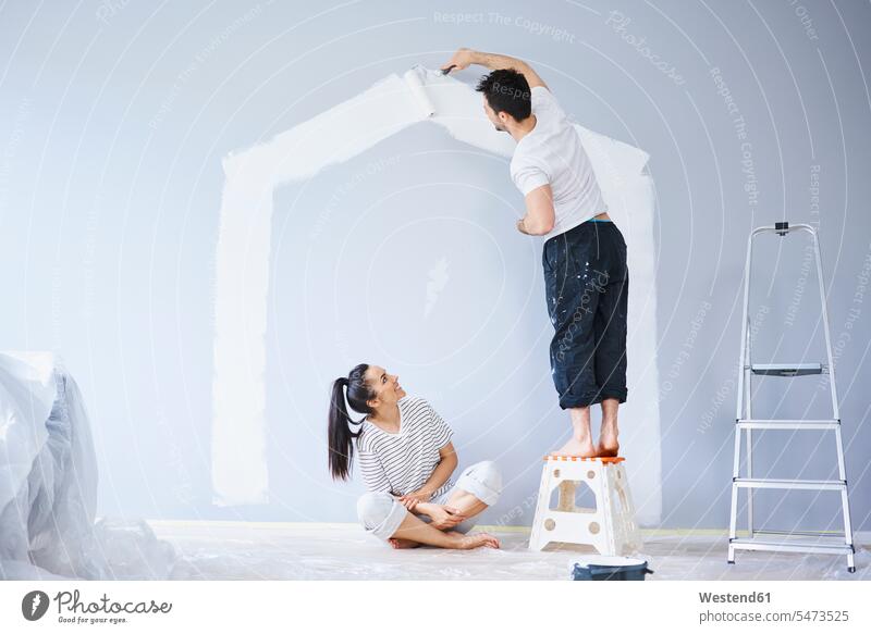 Ehepaar streicht Hausform an Wand in neuer Wohnung Form Formen wohnen Wohnungen Häuser Haeuser Wände Waende Paar Pärchen Paare Partnerschaft streichen Wohnen
