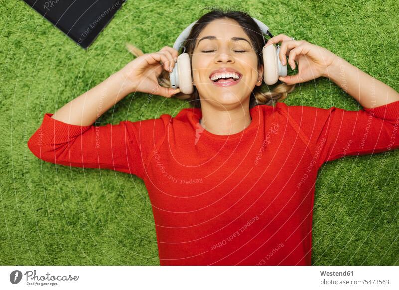 Glückliche junge Frau liegt auf einem Teppich und hört Musik mit Kopfhörern hören hoeren glücklich glücklich sein glücklichsein weiblich Frauen Teppiche liegen