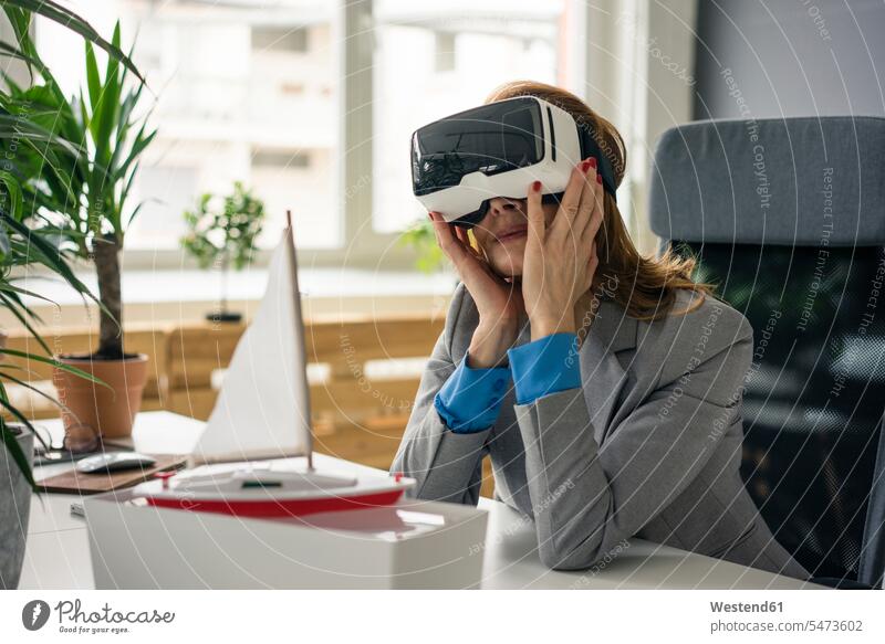 Geschäftsfrau sitzt am Schreibtisch mit einem Schiffsmodell, schaut durch eine VR-Brille Virtual Reality Brille Virtual-Reality-Brille Virtual Reality-Brille