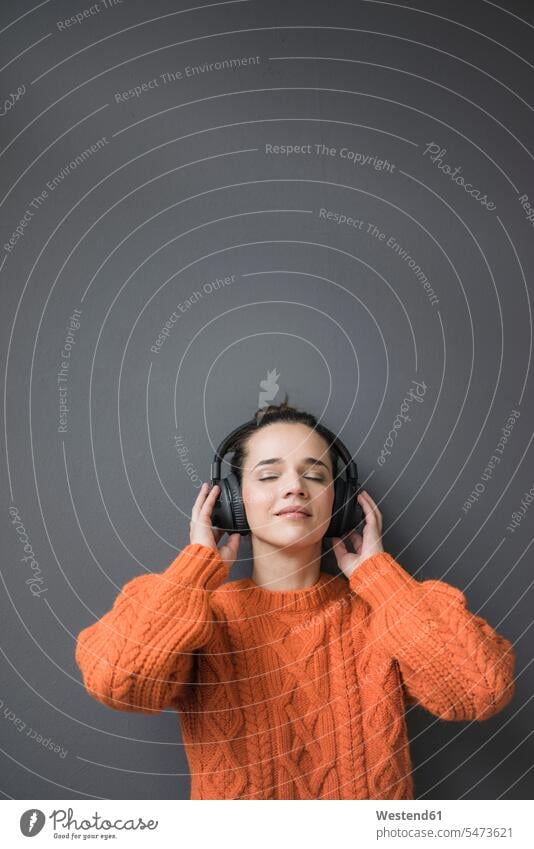 Porträt einer Frau mit orangefarbenem Strickpullover, die an einer grauen Wand lehnt und mit Kopfhörern Musik hört anlehnen angelehnt lehnend weiblich Frauen
