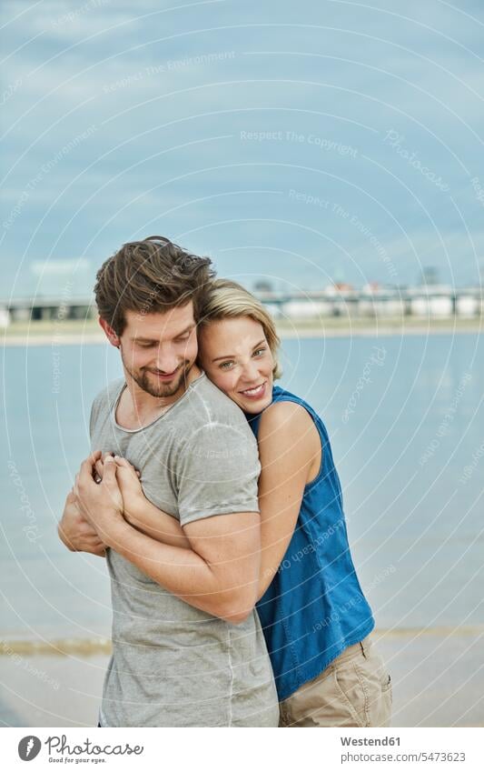 Glückliches junges Paar umarmt am Flussufer Fluesse Fluß Flüsse knuddeln umarmen Pärchen Paare Partnerschaft glücklich glücklich sein glücklichsein Gewässer