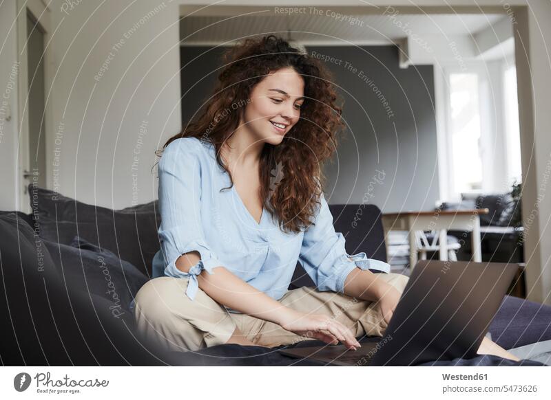 Junge Frau benutzt Laptop, während sie zu Hause auf dem Sofa sitzt Farbaufnahme Farbe Farbfoto Farbphoto Innenaufnahme Innenaufnahmen innen drinnen Tag