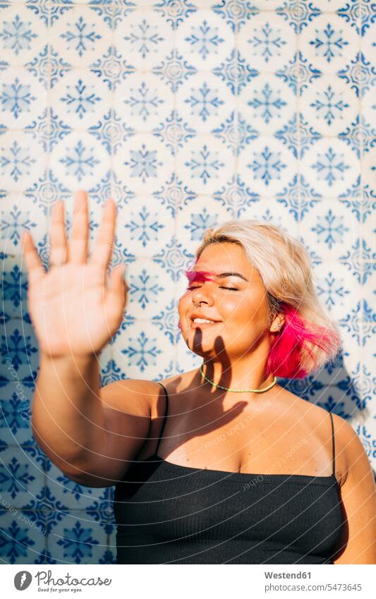 Lächelnde Frau blockiert Sonnenlicht mit der Hand an der Wand Farbaufnahme Farbe Farbfoto Farbphoto Außenaufnahme außen draußen im Freien Tag Tageslichtaufnahme