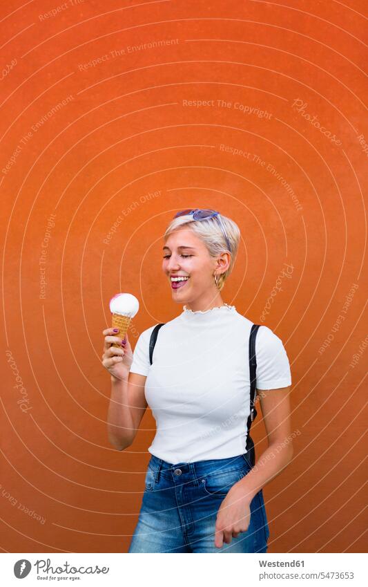 Lachende junge Frau mit Eiswaffel vor orangefarbenem Hintergrund lachen Eistüte Eistuete weiblich Frauen positiv Emotion Gefühl Empfindung Emotionen Gefühle