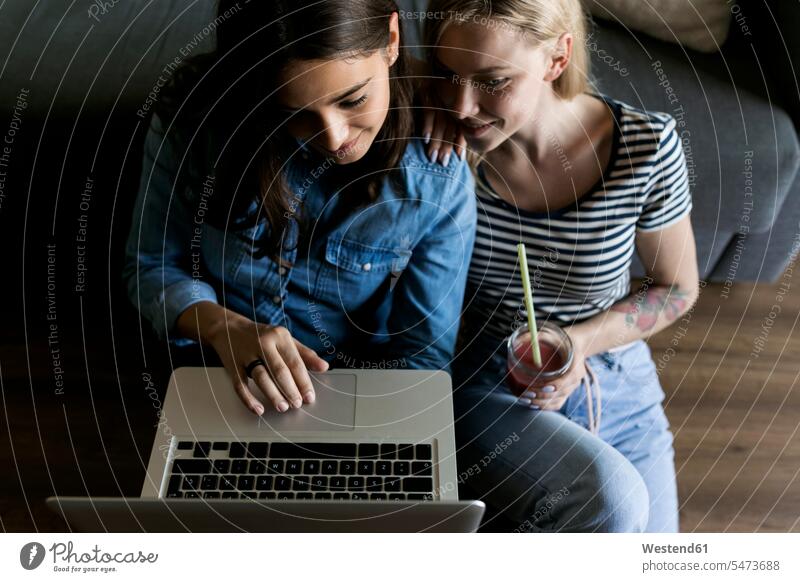 Zwei lächelnde junge Frauen sitzen auf dem Boden mit Softdrink teilen Laptop Notebook Laptops Notebooks Erfrischungsgetränk Erfrischungsgetraenk Freundinnen