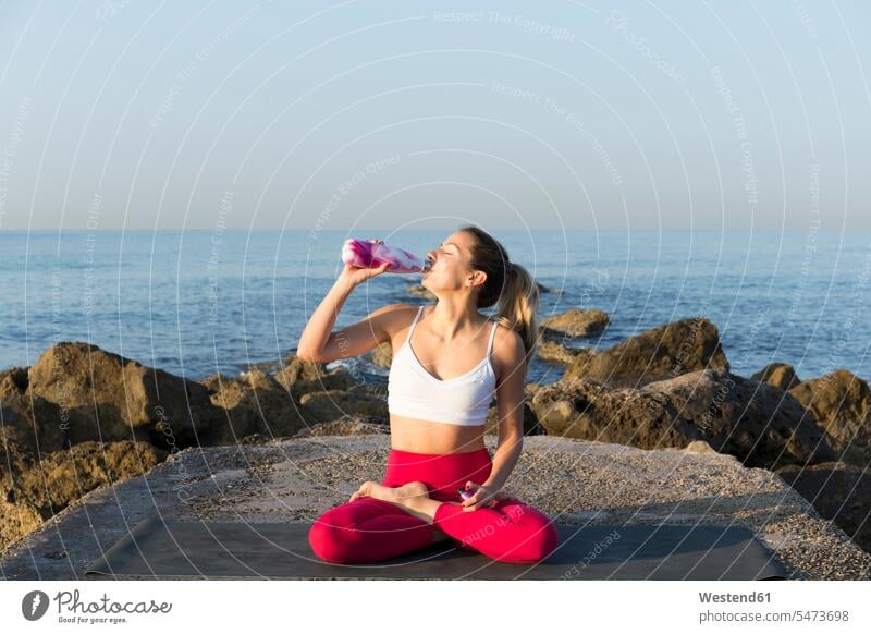 Junge Frau übt Yoga am Strand und trinkt Wasser sitzend sitzt ausüben trainieren Übung früh Frühe Morgen erfrischen erfrischend Beach Beaches Straende Strände