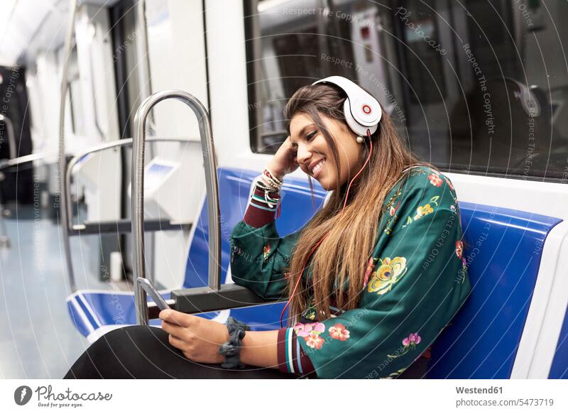 Lächelnde junge Frau hört Musik mit Kopfhörern und Smartphone in der U-Bahn lächeln iPhone Smartphones Kopfhoerer hören hoeren Ubahn U-Bahnen Untergrundbahnen