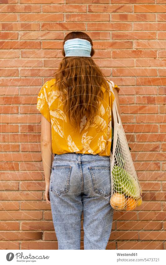 Junge Frau mit Lebensmitteleinkaufstasche mit Gesichtsmaske auf dem Haar, die vor einer Ziegelmauer steht Farbaufnahme Farbe Farbfoto Farbphoto Spanien