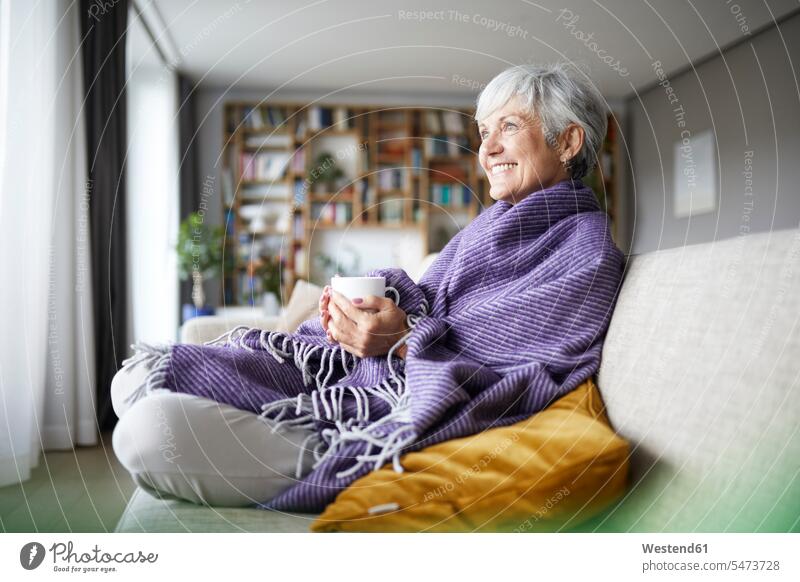 Lächelnde ältere Frau mit einer Decke, die eine Kaffeetasse hält, während sie zu Hause auf dem Sofa sitzt Farbaufnahme Farbe Farbfoto Farbphoto Innenaufnahme