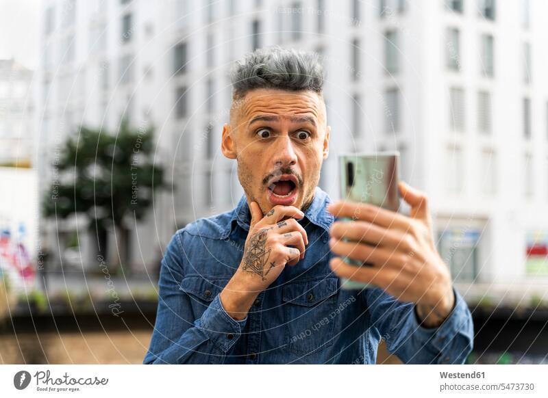 Schockierter homosexueller Mann hält Smartphone während Videoanruf gegen Gebäude Farbaufnahme Farbe Farbfoto Farbphoto Spanien Außenaufnahme außen draußen