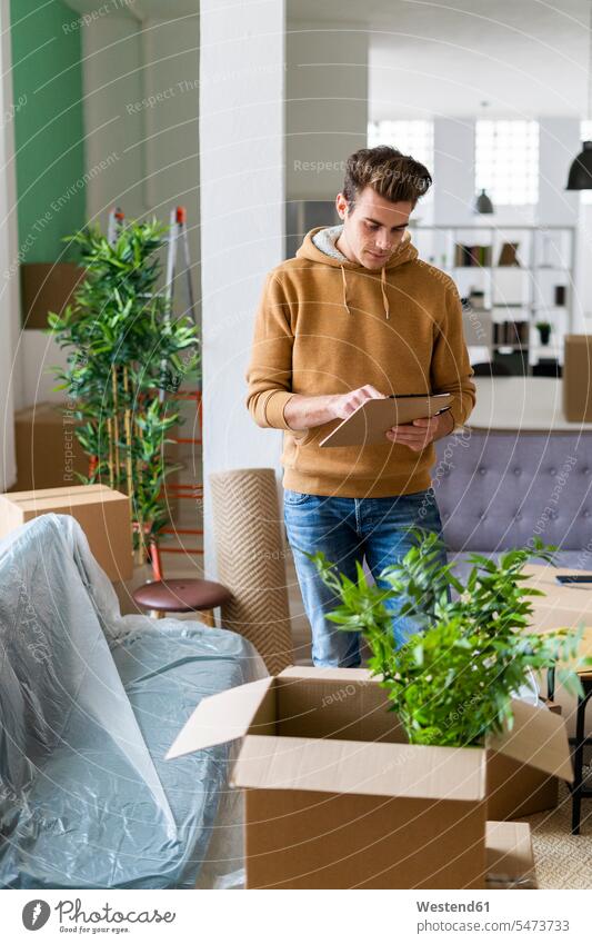 Junger Mann, der eine Liste hält, während er neben einer Kiste mit Pflanzen in einer neuen Loft-Wohnung steht Farbaufnahme Farbe Farbfoto Farbphoto