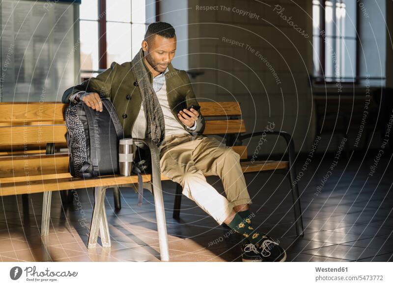 Stylischer Mann benutzt Smartphone, während er auf einer Bank in einem Bahnhof sitzt geschäftlich Geschäftsleben Geschäftswelt Geschäftsperson Geschäftspersonen