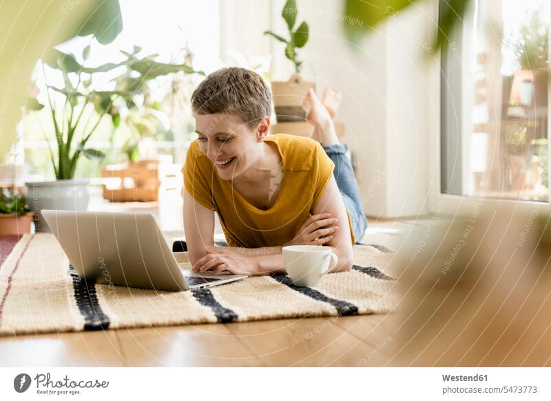 Lächelnde Frau mittleren Alters, die einen Laptop benutzt, während sie zu Hause auf dem Teppich liegt Farbaufnahme Farbe Farbfoto Farbphoto Deutschland