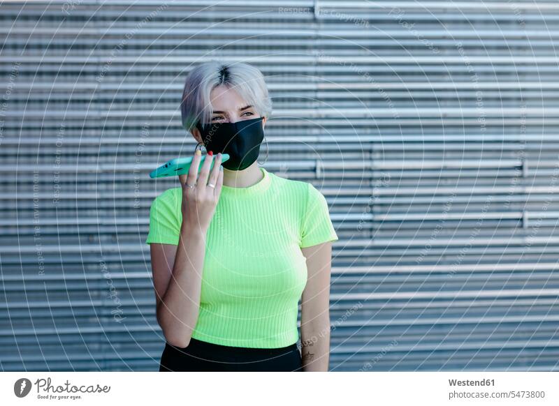 Blonde Frau steht vor einem Metallhintergrund, trägt eine Gesichtsmaske und benutzt ein Smartphone Metalle metallisch Telekommunikation telefonieren Handies