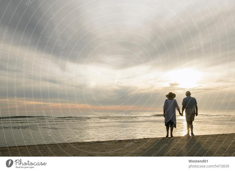Rückansicht des älteren Paares, das Hand in Hand am Strand steht und den Sonnenuntergang beobachtet, Liepaja, Lettland Leute Menschen People Person Personen