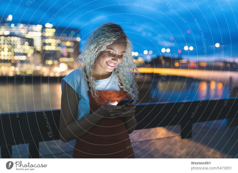Junge Frau in der Stadt in der Abenddämmerung mit Blick auf ihr Smartphone Informationen Telekommunikation telefonieren Handies Handys Mobiltelefon