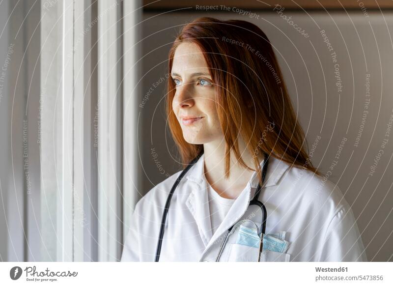 Lächelnde junge Ärztin schaut durchs Fenster Farbaufnahme Farbe Farbfoto Farbphoto Ärztinnen Doktorin Doktorinnen Arzt Ärzte Doktoren Medizin medizinisch